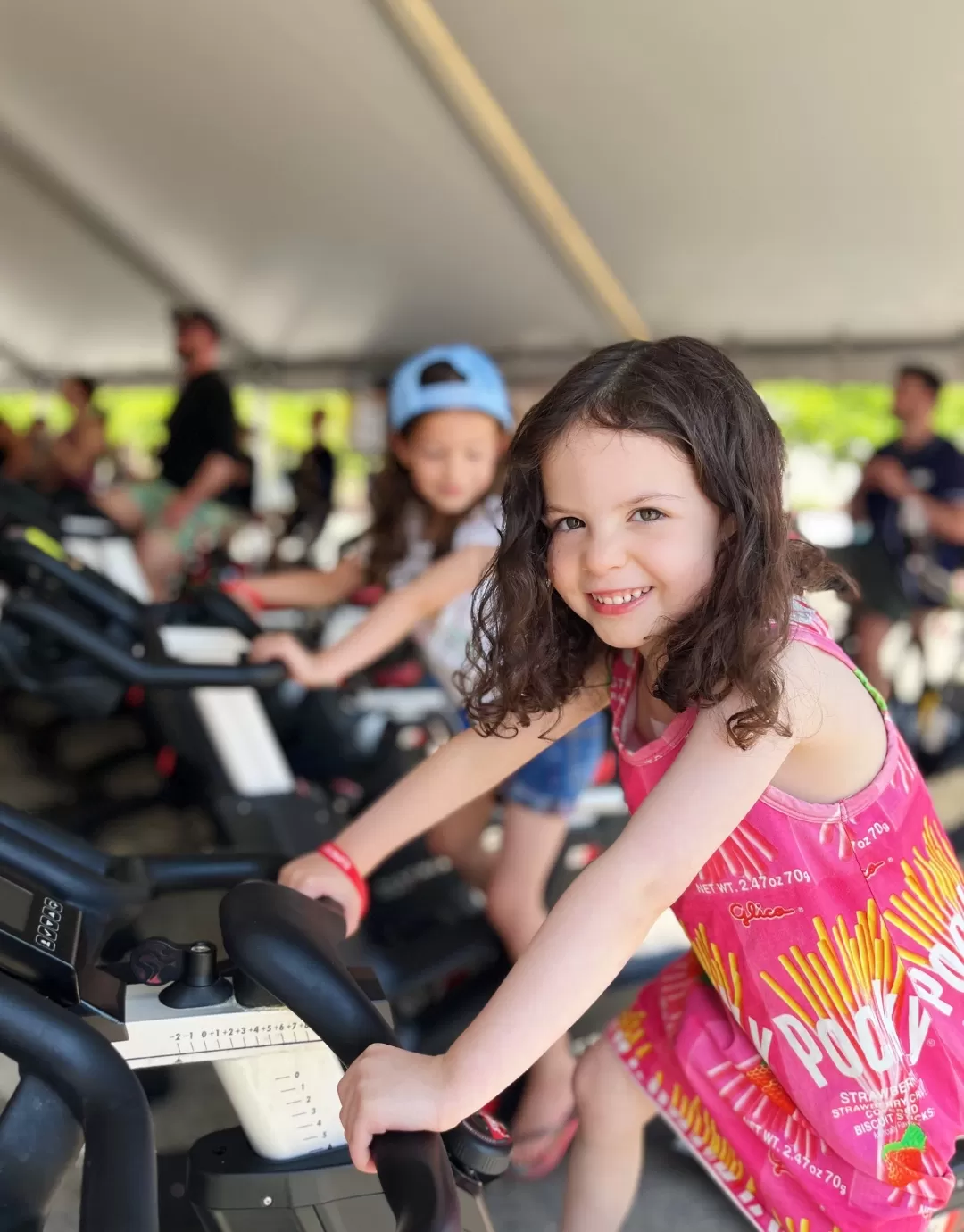 A 5 year old girl with long brown hair is sitting on a stationary cycle bike for adults and smiling at the camera. Behind her an outdoor cycle class is taking place under a big tent.