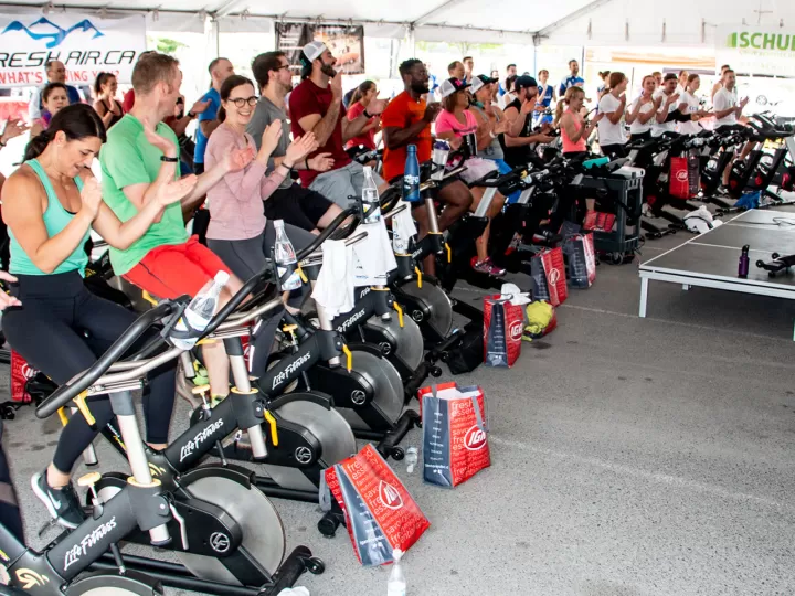 Large outdoor Cycle Class taking place under a tent with roughly 50 brightly dressed cyclists smiling and cheering