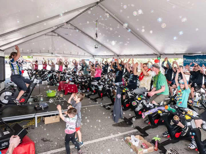  An outdoor Cycle class is under a large tent with dozens of participants dressed up brightly and cheering. There are bubbles blowing around the class and two children dancing in the foreground with one Cycle instructor on the a stage to the left leading the class. 