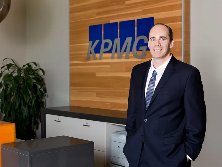 A professional shot of a man in a suite with light blue tie standing with his hands in his pockets smiling at the camera. There is a receptionist desk behind him with a sign that reads KPMG.