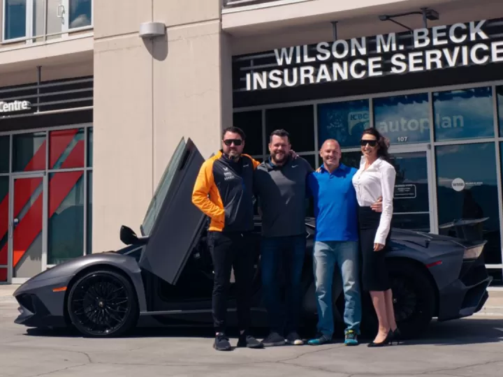 Three men and one woman are standing in front of a high-end grey sports car with suicide doors open. They have their arms around one another and are smiling at the camera. The photo is taken outside on a sunny day with offices behind them. 