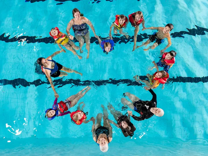 15 community members of all ages from senior to child, holding hands in a pool and looking up at the camera. Their bodies are forming the shape of a heart. 